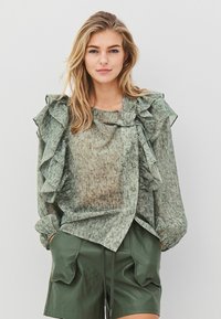 Ljusgrön blus med mönster, rynkade detaljer och knappar fram, i kombination med mörkgröna shorts.