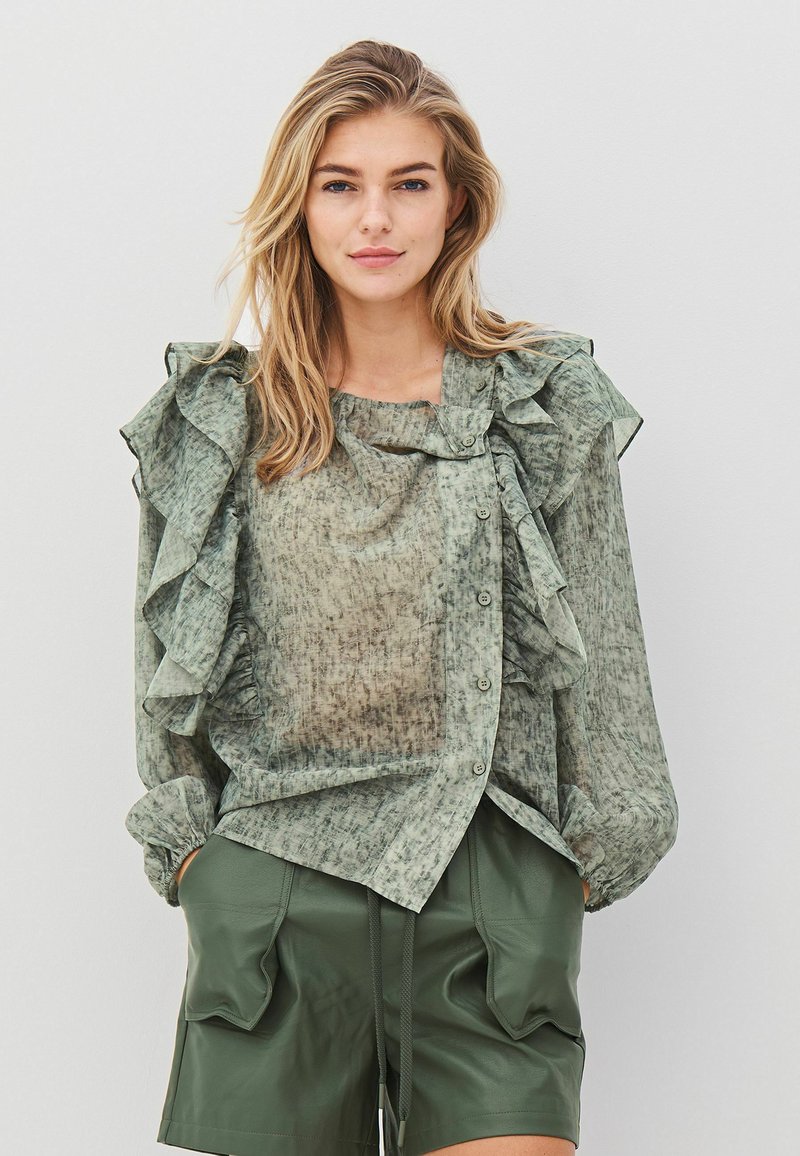 Ljusgrön blus med mönster, rynkade detaljer och knappar fram, i kombination med mörkgröna shorts.