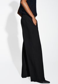 Pantalons larges noirs en tissu lisse, dotés de poches latérales et d'une taille ajustée, accentués par un design élégant et une silhouette longue.