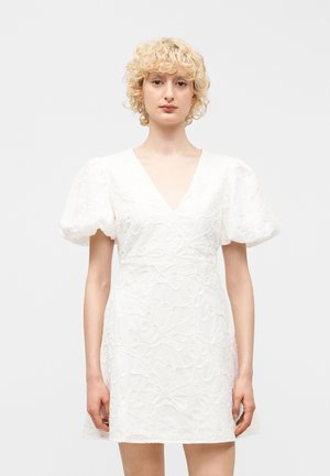 Frau mit kurzen, lockigen blonden Haaren, die ein weißes, strukturiertes Kleid mit gepufften kurzen Ärmeln und tiefem V-Ausschnitt trägt, vor einem schlichten Hintergrund stehend.