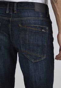 Tamsiai mėlyni džinsai su siauru kirpimu, su geltonais siūlais, užpakalinėmis kišenėmis ir odine etiketėje ant juosmens.