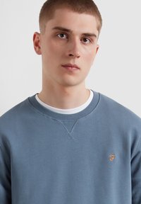 Felpa in cotone blu con scollo rotondo, caratterizzata da un piccolo logo ricamato arancione sul petto e polsini e orlo a costine.