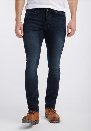 Man met donkerblauwe slim-fit jeans en bruine leren veterlaarzen, staand tegen een witte achtergrond.