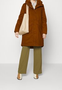 Brun parka med en pälskantad huva, justerbara snoddar i midjan, beige tote-väska, gröna vida byxor och tvåtonade klackstövlar.