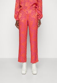 Pantalons à motifs roses et oranges avec un design fluide, fabriqués en tissu doux. Présentent une jambe large et des poches latérales. Les chaussures sont de style plateforme blanche.