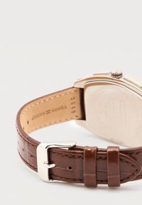 Καφέ δερμάτινο λουράκι ρολογιού με ασημένια αγκράφα, ραμμένα άκρα και το όνομα της μάρκας χαραγμένο στο εσωτερικό του λουριού κοντά στη θήκη του ρολογιού.