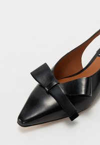 Zapato de cuero negro de punta con acento de lazo, talón abierto y forro interior en color beige. Textura suave con un acabado pulido.