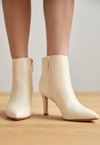 Bottines couleur crème à bout pointu avec fermetures éclair latérales et talons fins, portées sur jambes nues, debout sur un sol en bois.