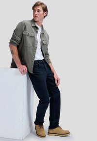 Ung mand, der læner sig op ad en hvid blok, iført khaki jakke, hvid skjorte, mørke jeans og beige støvler, ser svagt mod venstre.