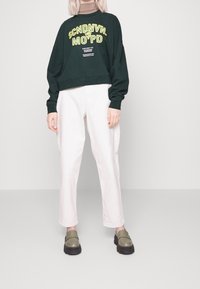 Felpa verde cropped con testo giallo audace, pantaloni beige a gamba larga e scarpe slip-on verde oliva con suole nere spesse.