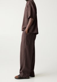 Osoba stojąca bokiem, ubrana w luźne, ciemnobrązowe lniane spodnie, pasującą koszulę z krótkim rękawem i ciemnobrązowe sandały na otwartym palcu z klamerką.