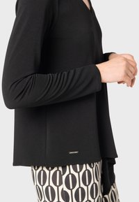 Svart långärmad topp med V-ringning, med en avslappnad passform och gathered ärmar, tillsammans med mönstrade byxor. Metallbackning i fållen.