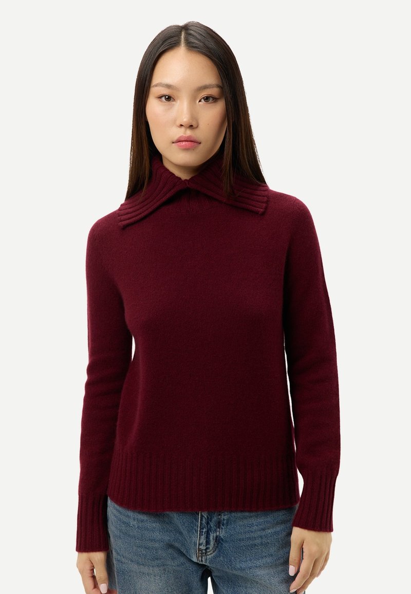 Femme aux cheveux longs et raides portant un pull bordeaux à col large côtelé et un jean bleu, debout devant un fond uni.