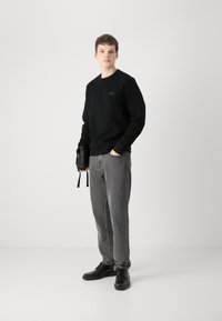 Crna dukserica s izvezenim logotipom, sivi traperice i crne cipele. Model drži malu crnu torbicu, postavljen protiv jednostavne pozadine.