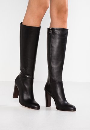 Černé kožené kozačky po kolena s vysokými širokými podpatky a bočními zipy, nošené na bosých nohách, zobrazené na jednobarevném bílém pozadí.