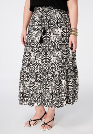 Vrouw die een zwart-witte bloemen midi-rok draagt met kwastjeskoord, zwarte top, zwarte sandalen met bandjes en meerdere armbanden aan één pols.