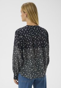 Woman with long blonde hair facing away wearing a black and white floral long-sleeve blouse and blue jeans against a white background.