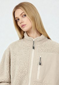 Giacca in pile beige con collo alto, chiusura con zip, due tasche con zip e superficie testurizzata. Dettagli includono tiretti delle zip neri.