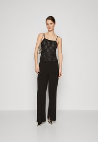 Haut sans manches noir avec des clous métalliques, associé à un pantalon noir taille haute. Le modèle tient un sac à main en argent scintillant et porte des talons noirs.
