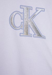 Tela de color púrpura claro con un logo "CK" plateado texturizado, contorneado por un fino borde azul claro. El diseño es audaz y moderno.