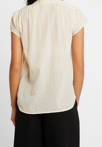 Blouse beige clair avec de fines rayures verticales, manches courtes et ourlet arrondi. Tissu texturé avec de subtils plis près du col.