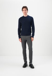 Dunkelblaues strukturiertes Strickpullover mit rundem Ausschnitt, kombiniert mit schmalen grauen Hosen und schwarzen Slippern. Das Model steht vor einem weißen Hintergrund.