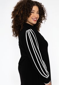 Chemise noire avec un col à boutons, ornée de rayures blanches sur les manches. Le tissu semble lisse et offre une coupe décontractée.