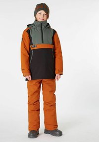 Orange und schwarze Skijacke mit einem grünen Brustpanel, kombiniert mit passenden oran fillen Hose und grauen Stiefeln. Beinhaltet eine schwarze Mütze mit orangefarbener Schrift.
