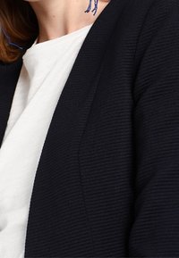 Černý žebrovaný blazer s texturovaným finišem, nošený přes bílý oděv. Má hluboký výstřih do V a šikmé klopy, což vytváří upravený vzhled.