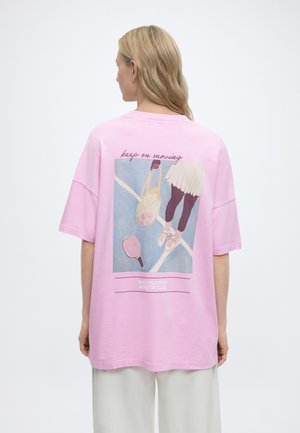 Hosszú szőke hajú nő, aki egy rózsaszín, túlméretezett pólót visel teniszes témájú grafikával és a hátoldalon a "keep on moving" felirattal.