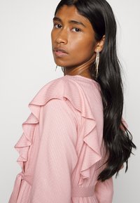 Vestido rosa com babados, apresentando um tecido texturizado, mangas longas e uma cintura atada. A modelo usa brincos prateados longos e assimétricos.