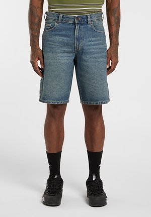 Pantalones cortos de mezclilla azul con corte recto, cinco bolsillos y cierre de botón. Combinados con zapatillas deportivas negras y calcetines.