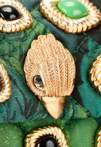 Spilla a forma di uccello in oro con dettagli a foglia, caratterizzata da un occhio in pietra preziosa trasparente, posizionata su uno sfondo di tessuto verde trapuntato con accenti neri e verdi.