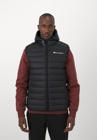 Chaleco acolchado negro con capucha, que presenta secciones acolchadas horizontales y una cremallera frontal. Logotipo de Champion mostrado en el lado izquierdo del pecho.