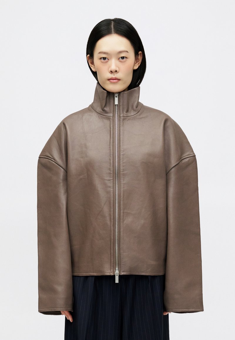 Καφέ δερμάτινο μπουφάν με ψηλό γιακά, φαρδιά γραμμή, μπροστινό φερμουάρ και φαρδιές μανίκες. Η υφή είναι λεία με ματ φινίρισμα.