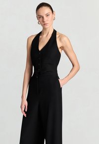 Donna con top nero senza maniche a collo ad anello e pantaloni a gamba larga a vita alta, in piedi con una mano in tasca contro uno sfondo chiaro.