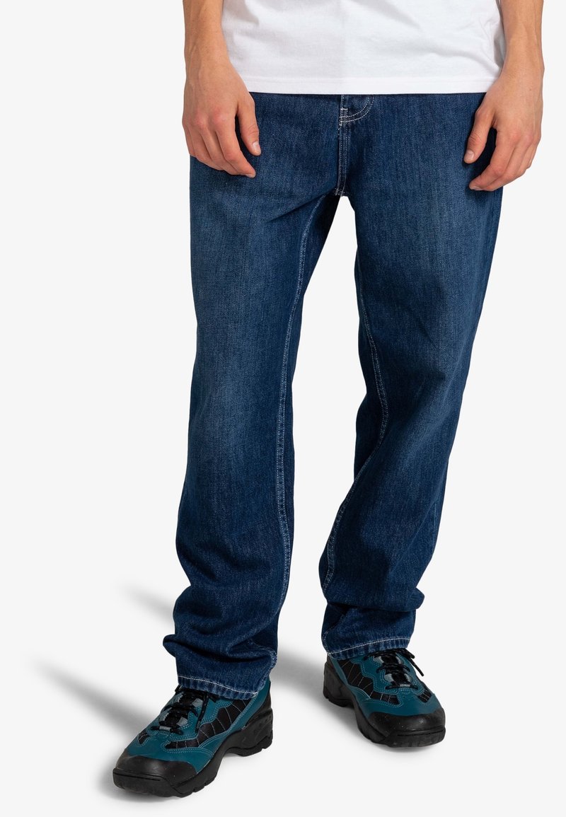 Jean en denim bleu foncé avec une coupe droite, présentant une disposition classique à cinq poches, associé à des chaussures à lacets noires et bleu sarcelle.