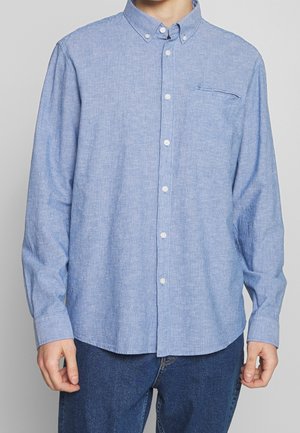 Mężczyzna w jasnoniebieskiej, w paski, zapinanej na guziki koszuli z długim rękawem i zapinanym kołnierzykiem oraz ciemnoniebieskich dżinsach, stojący na neutralnym tle.