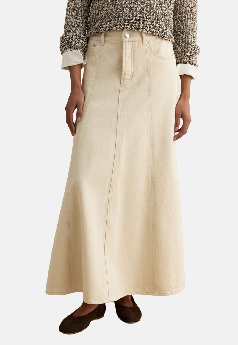 Mujer con falda vaquera beige en línea A con detalles de costuras frontales, combinada con un suéter marrón texturizado y zapatos planos marrón oscuro.