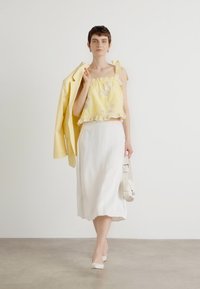 Un crop top jaune à volants avec un motif floral, associé à une jupe midi en satin blanche. Le mannequin tient un blazer jaune clair et un sac à main blanc.