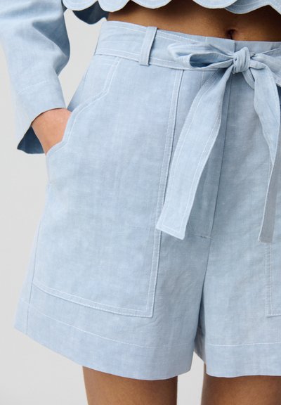 Pantalones cortos de lino azul claro con cintura ajustable, que cuentan con bolsillos laterales y una textura suave, terminados con costuras de contrasta blancas.