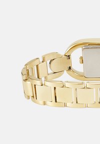 Goldfarbene Uhr mit einem geometrischen Gliederarmband, das ein rechteckiges Gehäuse und eine glatte Oberfläche aufweist. Einfarbig, polierte Metalloberfläche.