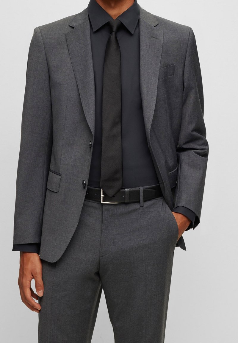 Homme portant un costume gris foncé avec une chemise noire, une cravate noire et une ceinture noire, debout avec une main dans la poche devant un fond uni.