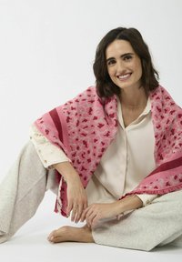 Roze patroon sjaal met franjes, over een lichte blouse gedrapeerd. Draagt over een neutraal gekleurde outfit, met verschillende tinten roze.