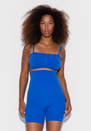 Frau mit lockigem Haar trägt ein leuchtend blaues, figurbetontes zweiteiliges Sportoutfit vor einem schlichten weißen Hintergrund.