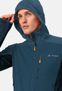 Giacca da uomo Vaude di colore blu scuro, con finitura testurizzata, cappuccio regolabile e dettagli arancioni sulla zip. Due tasche con zip sul davanti.