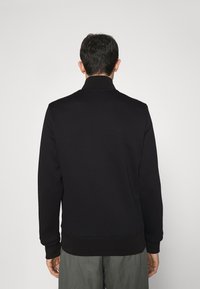 Homme portant une veste noire à manches longues et un pantalon gris, se tenant de dos face à l'appareil photo sur un fond clair et uni.