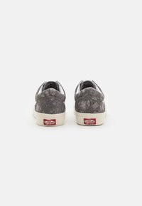 Zapatillas deportivas grises con un acabado texturizado, acentos blancos y suelas de goma. Presentan un logo de Vans rojo en el talón.