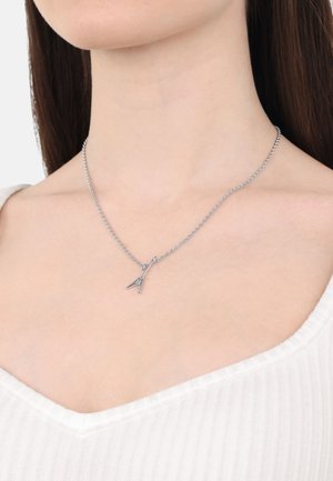 Ciondolo d'argento con una catena sottile e un pendente affusolato, appoggiato sulla pelle vicino alla clavicola, che completa un top bianco.
