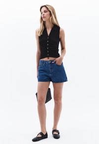Svart ärmlös knäppt topp med rynkade detaljer, kombinerad med denim-shorts och svarta flats. Modellen håller en texturerad svart kuvertväska.